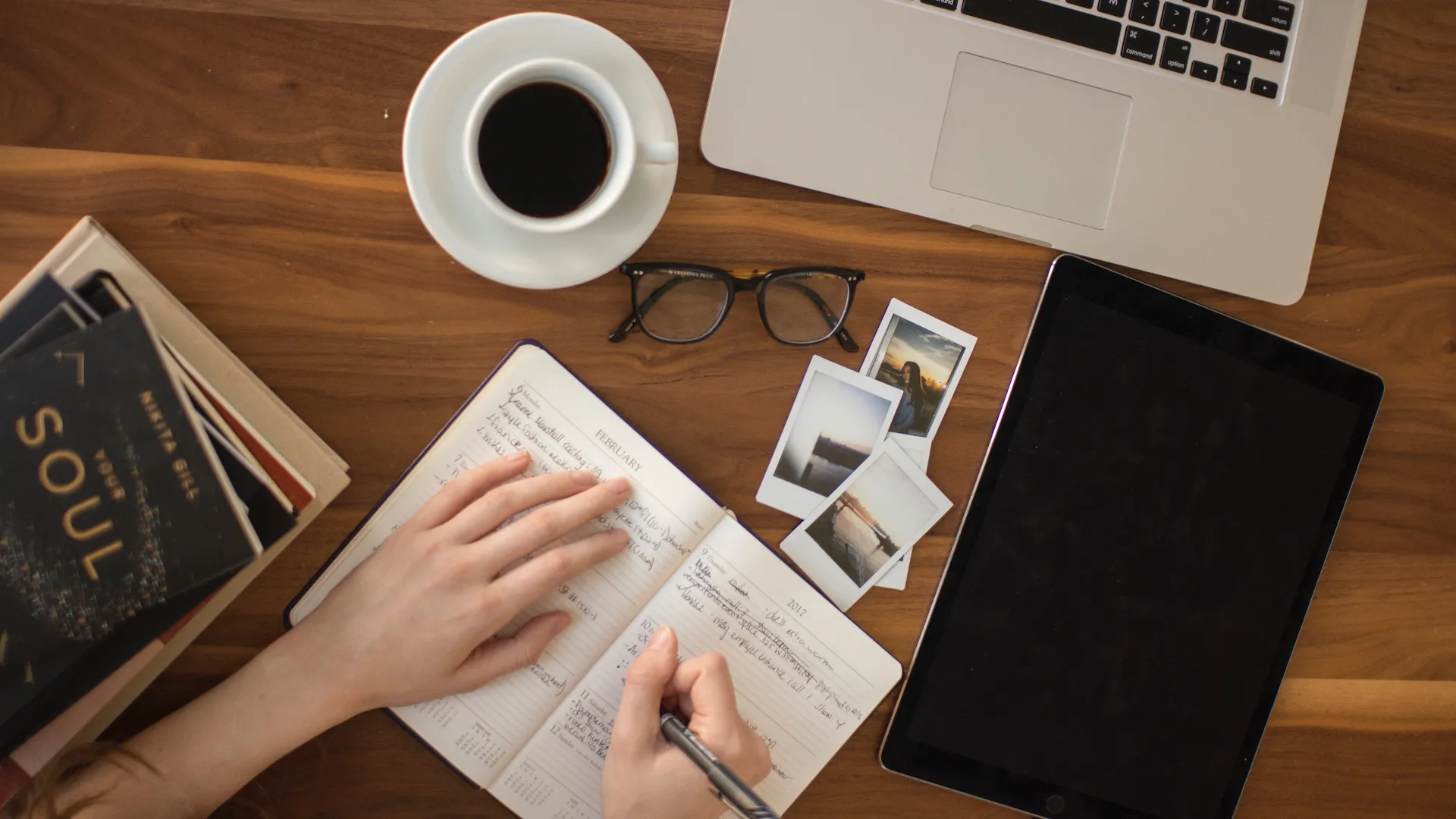 Human writing notes on a table with books, coffee, pictures, glasses and a laptop